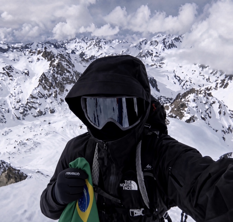 Rafael Bueno in ski goggles and a black jacket, holding a Brazilian flag in snowy mountains